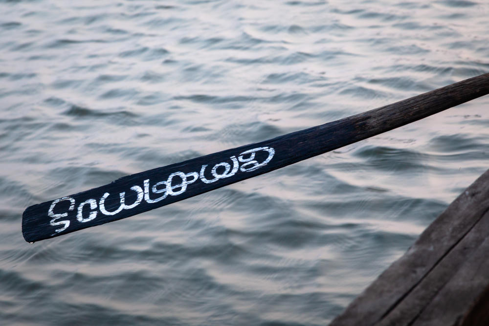 A wooden boat oar with a dark blade hand-painted in white with text in a South Asian script, held out over the calm gray water of the Kaithappuzha Backwaters, with the worn edge of a wooden boat visible in the lower right.