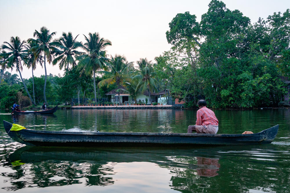 A man in a pink checked shirt sits in a long, narrow wooden canoe on calm green backwaters, with a second small boat carrying people in the distance. The far shore is lined with coconut palms and dense greenery, with a small white house among the trees under a pale evening sky.