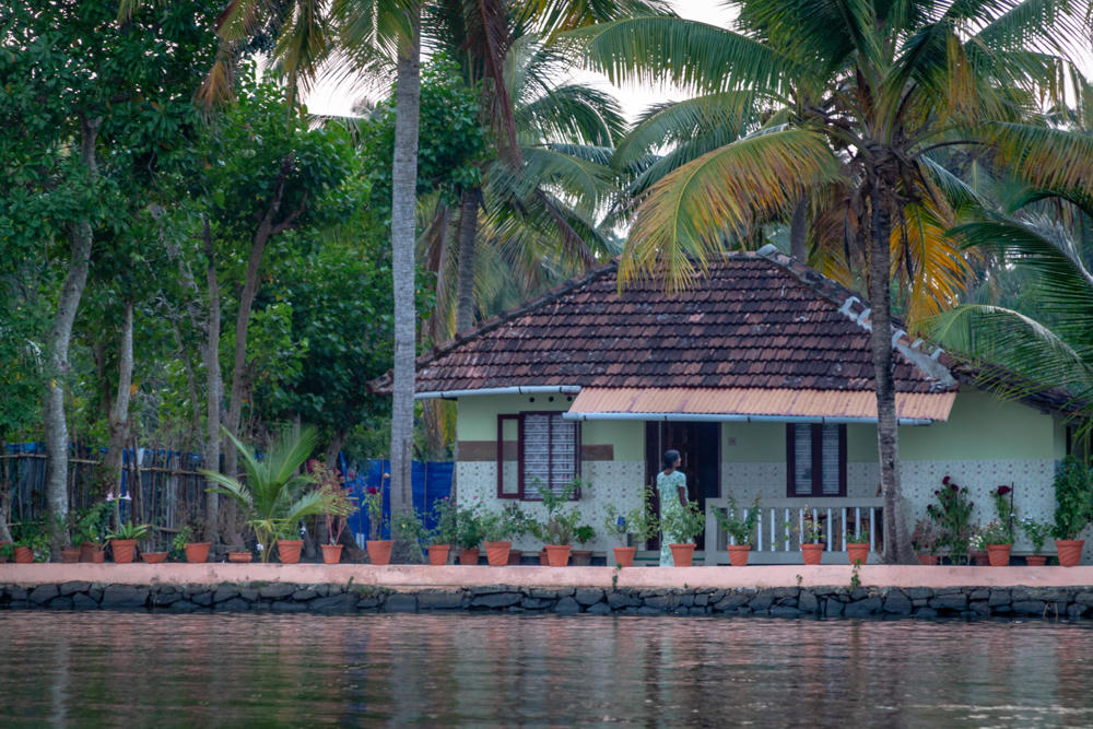 A traditional tiled-roof house sits along the edge of the Kaithappuzha Backwaters, fronted by a row of terracotta potted plants on a paved walkway above a stone embankment, with a person in a light green outfit standing on the veranda. Tall coconut palms and lush green trees rise behind the house, and the calm backwater reflects the scene in the foreground.