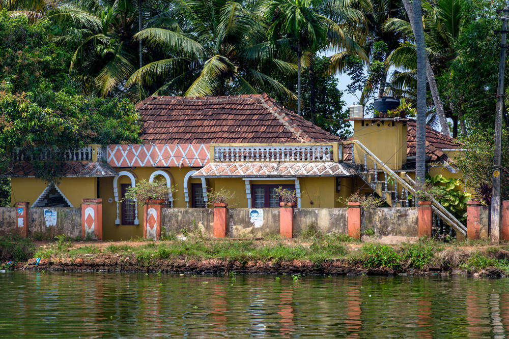 A yellow tile-roofed house with a decorative white balustrade and arched veranda sits behind a low compound wall with orange pillars along a green backwater, framed by tall coconut palms and dense tropical foliage under a bright sky.
