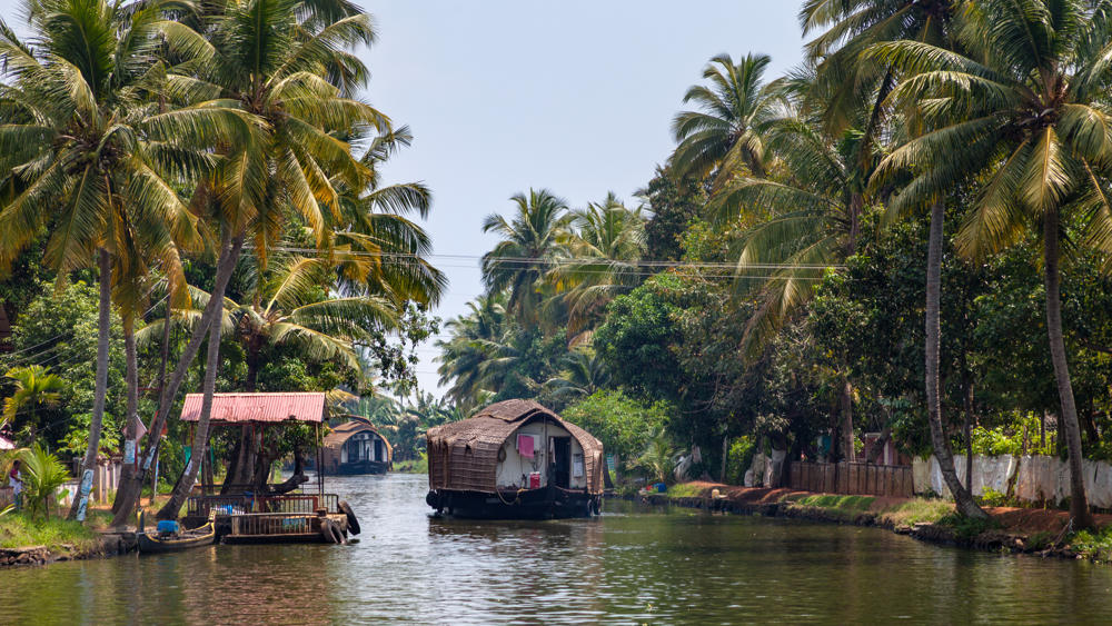 A traditional thatched-roof houseboat drifts along a calm green backwater channel lined with tall coconut palms and dense tropical foreground, with a covered dock and small dwellings on the left bank under a hazy midday sky.