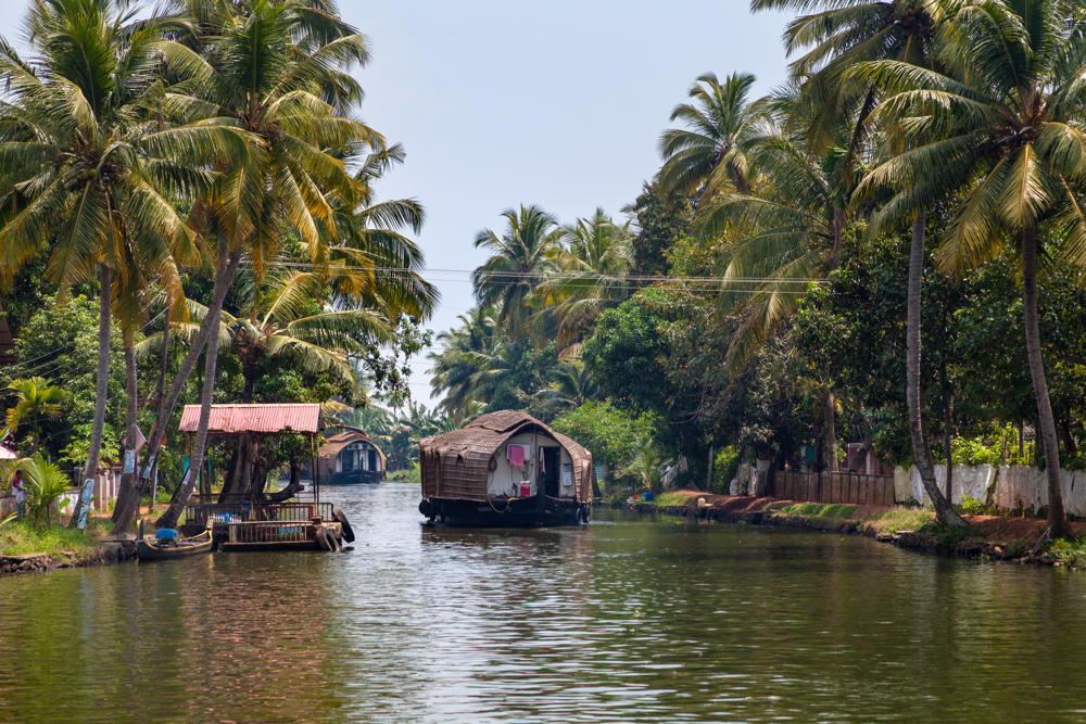 A traditional thatched-roof houseboat drifts along a calm green backwater channel lined with tall coconut palms and dense tropical foreground, with a covered dock and small dwellings on the left bank under a hazy midday sky.