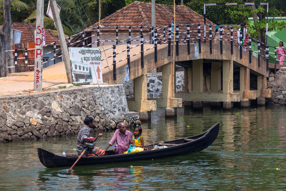 Three people – a man, another man, and a young girl in a yellow dress – travel in a narrow wooden canoe on green backwater, with one man rowing at the stern. Behind them a concrete arched footbridge with black-and-white striped railings crosses the water, alongside a stone embankment, tiled-roof buildings, palm trees, and signs in Malayalam.