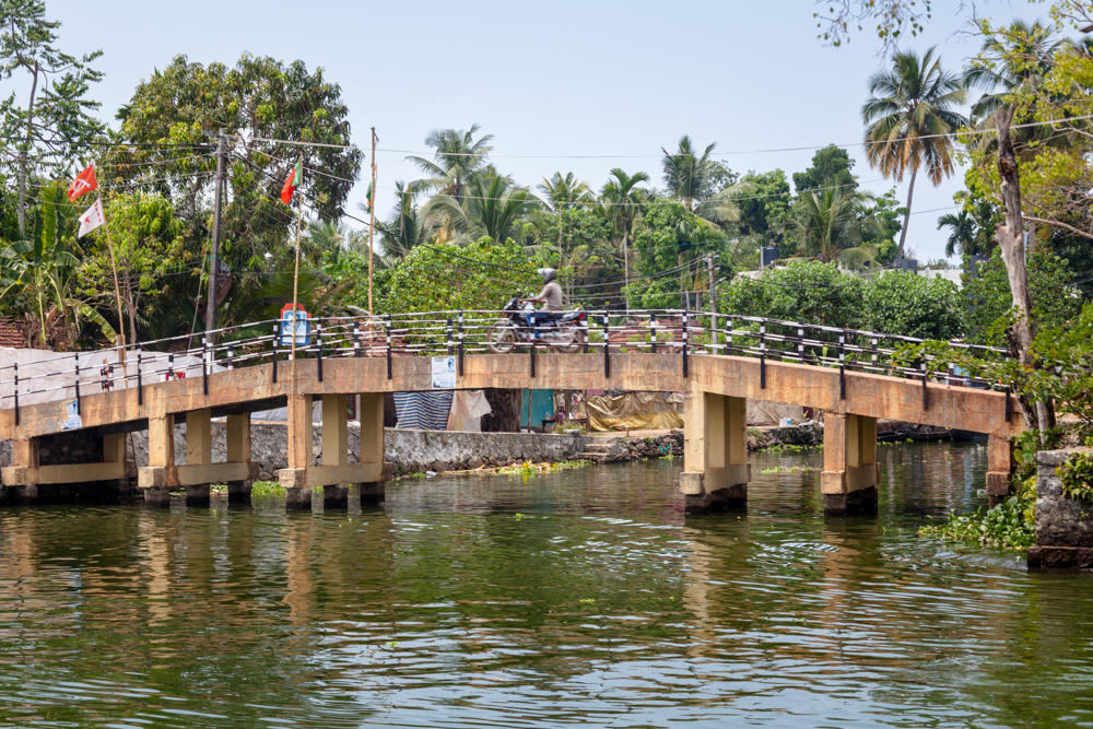 A person on a motorcycle crosses a low concrete arched bridge over a green waterway, with lush palm trees, tropical greenery and small red flags on poles lining the far bank.