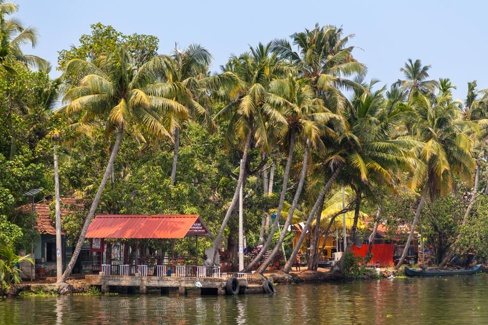 A palm-fringed backwater shoreline with tile-roofed and orange-roofed buildings along the water’s edge, a waterfront pavilion with white railings and old tires hung for mooring, and a narrow canoe pulled up to the bank under a hazy sky.