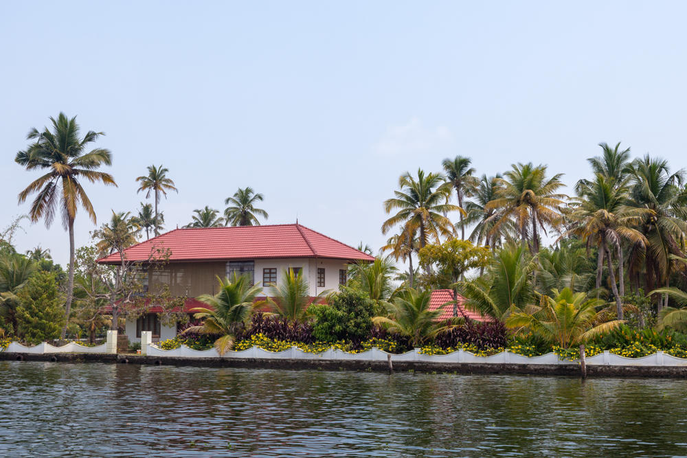 A white house with a red tiled roof stands on the banks of a waterway in Kerala, surrounded by tall coconut palms and lush greenery, with a low white wall and yellow flowers lining the water’s edge under a hazy sky.