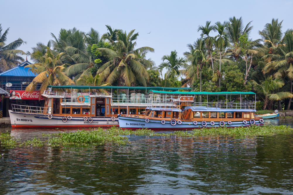 Two wooden passenger ferry boats named “St. Mary’s” and “St. Sebastian” moored side by side on a backwater channel lined with dense coconut palms, with water hyacinth floating on the greenish water and a waterside building bearing a Coca-Cola sign on the left.