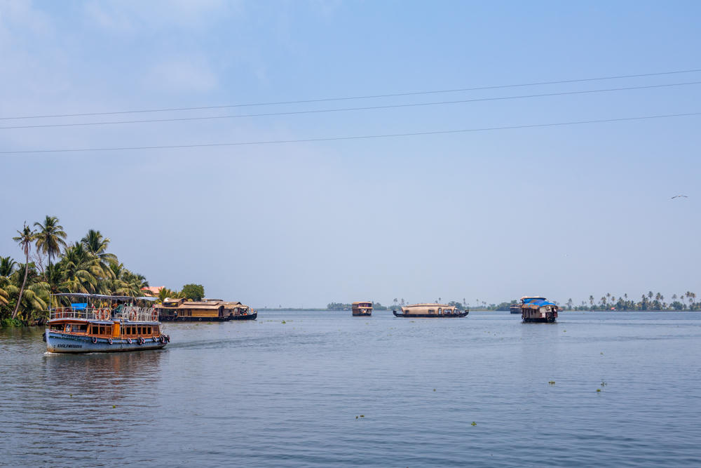 A wooden passenger ferry with a canopy cruises along a broad stretch of the Kerala backwaters, with several traditional houseboats scattered across the water in the distance. The left bank is lined with dense palm trees and low buildings under a hazy pale blue sky.