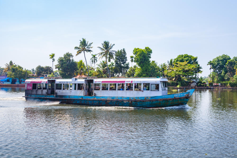 A weathered public passenger ferry with a white and pink cabin and a rusted blue-and-white hull motors along a broad river, with passengers seated inside behind open windows. Palm trees and dense green foliage line the far bank under a hazy pale sky.