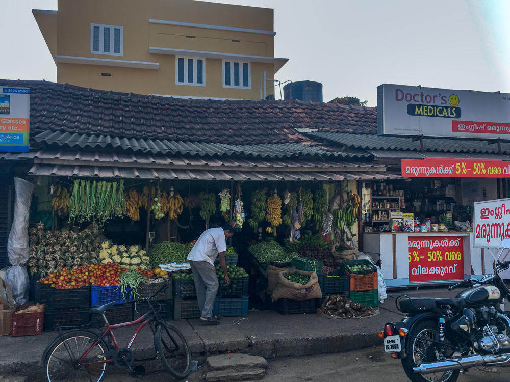A tile-roofed roadside fruit and vegetable stall in Alappuzha, Kerala, with hanging bunches of bananas and displays of pineapples, tomatoes, cauliflower, and green beans in crates, where a man in a white shirt bends over to arrange produce. A red bicycle and a black Royal Enfield motorcycle are parked out front, and signs in Malayalam and English, including “Doctor’s Medicals” and a Reliance Mobile ad, line the shopfronts.