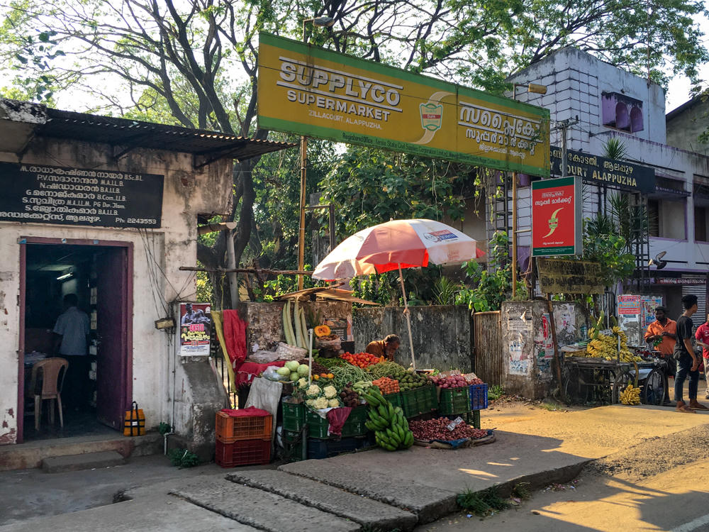 A street-side vegetable and fruit stall in Alappuzha, Kerala, with produce like bananas, cauliflower, tomatoes, mangoes and onions displayed in crates under a red-and-white umbrella, beneath a yellow and green Supplyco Supermarket sign written in English and Malayalam. A weathered building with a doorway sits at left, and people stand near a cart of bananas along a sunlit road at right.