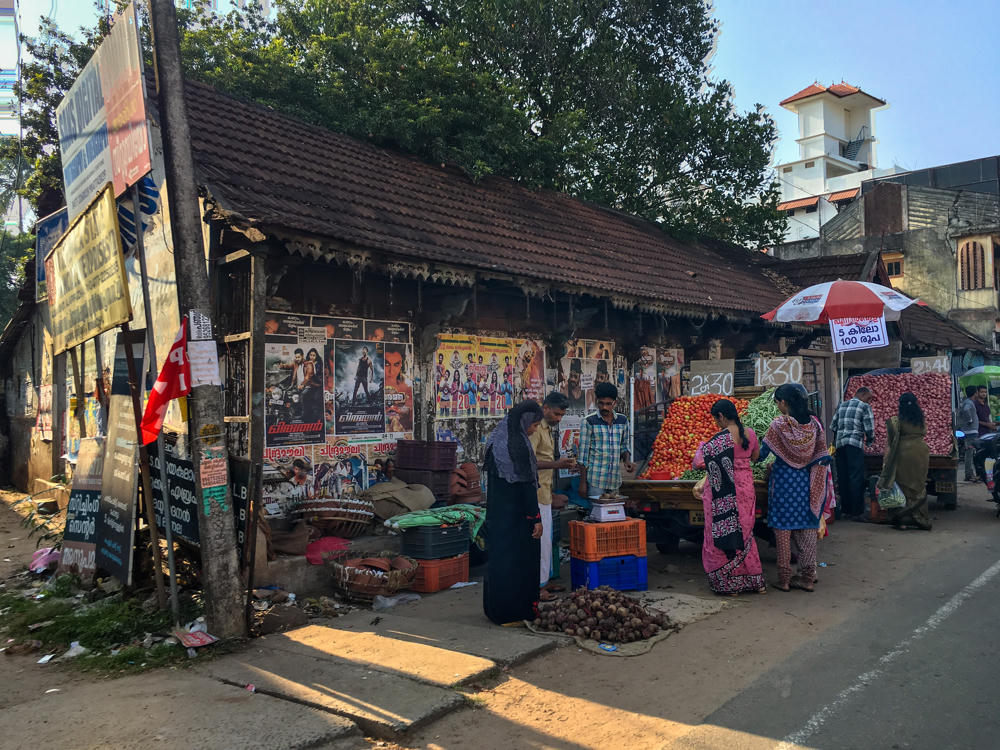 A busy roadside vegetable market in front of an old tiled-roof building plastered with colorful Malayalam film posters, where vendors sell tomatoes, green chilies, and onions from crates and carts. Several women in saris and salwar suits shop alongside a man in a plaid shirt weighing produce, with a red-and-white promotional umbrella and a white tower building visible in the background.