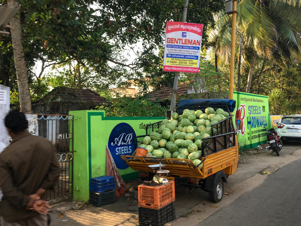 A yellow three-wheeled auto truck piled high with striped watermelons parked on a roadside in Alappuzha, Kerala, with a balance scale and stacked plastic crates beside it. Behind it are bright green painted walls advertising “A.R Steels” and “Steels & Hardwares,” a red Malayalam sign for Gentleman Chit Funds, palm trees, and a man in brown walking past in the foreground.