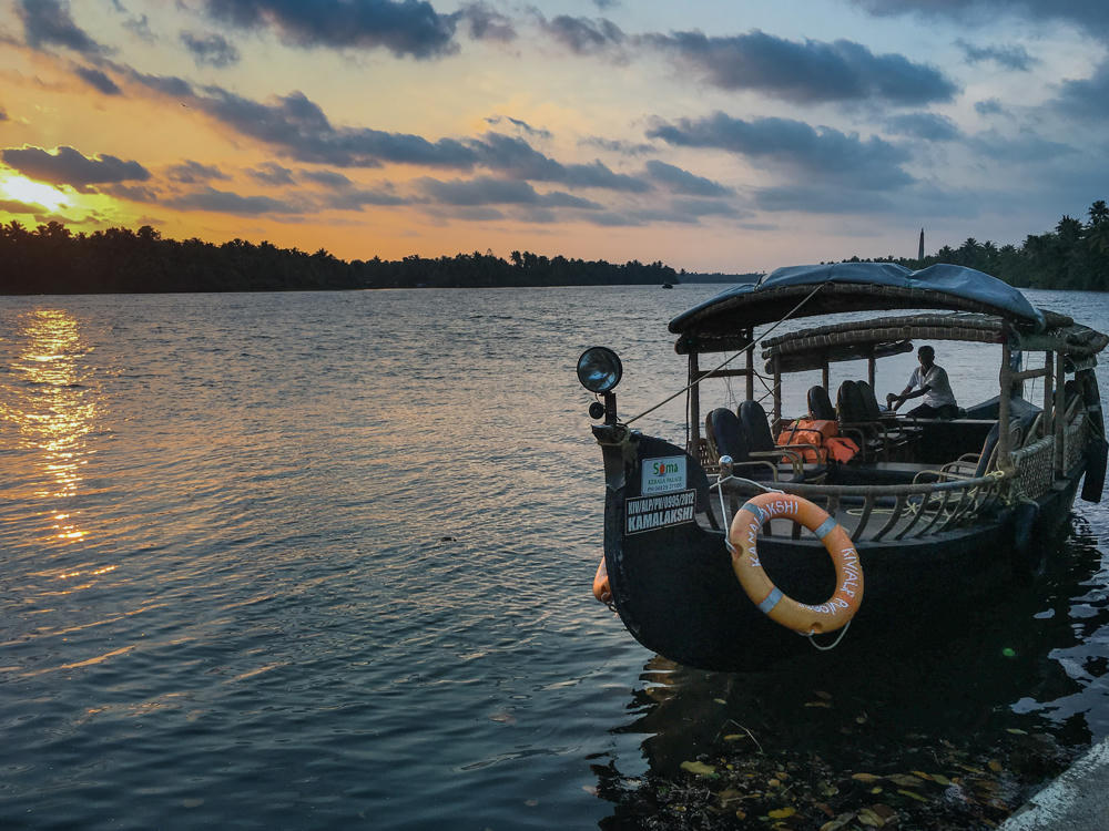 A small covered tour boat named “Kamalakshi” moored on calm backwaters at sunset, with a man seated at the helm and an orange life ring hanging on its bow. Palm trees line the far shore beneath a dramatic sky glowing orange and pink over the water.