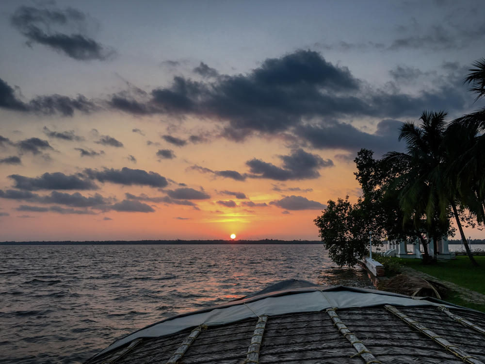 A sunset over calm backwaters seen from the curved thatched-and-canvas roof of a houseboat, with the orange sun low on the horizon beneath a sky of scattered grey clouds. On the right, silhouetted palm trees and greenery line the shore near a white columned building.