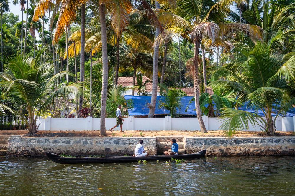 Two men in a narrow wooden canoe on the Manimala River backwaters, one paddling near a low stone embankment lined with a white wall and dense coconut palms, while a third man in a lungi walks along the shore path behind them.