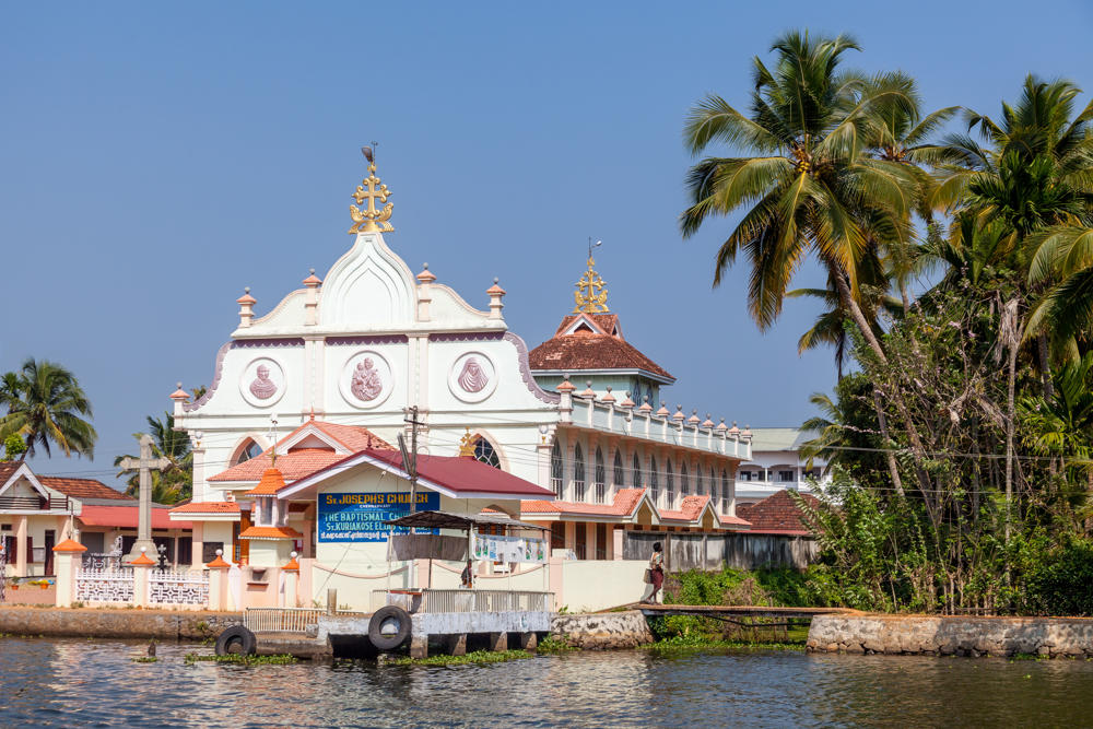 A white church with an ornate gabled facade topped by golden crosses stands along the edge of the Pamba River, with a blue sign reading “St. Joseph’s Church” near its entrance and a small waterside jetty in front. Tall coconut palms rise beside the building against a clear blue sky, seen from across the water in the Kerala backwaters.