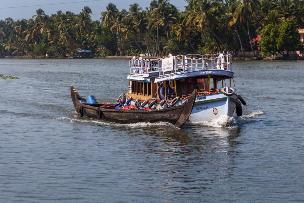 A double-decker passenger ferry towing a long wooden canoe laden with cargo travels along the calm backwaters of the Pamba River, with a dense line of coconut palms and a few buildings along the far bank under a hazy sky.