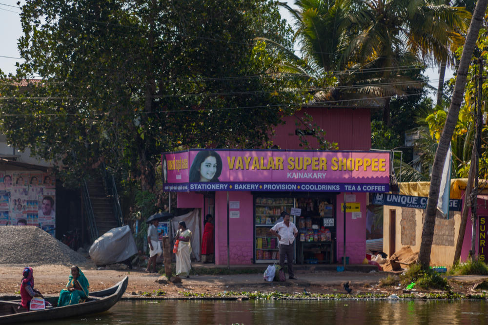 A pink roadside shop called “Vayalar Super Shoppee” in Kainakary, Kerala, selling stationery, bakery, provisions, cool drinks and tea and coffee, seen from across a canal where two women in colorful saris sit in a wooden canoe. People stand near the shop under tall palm trees and dense green foliage in the midday sun.