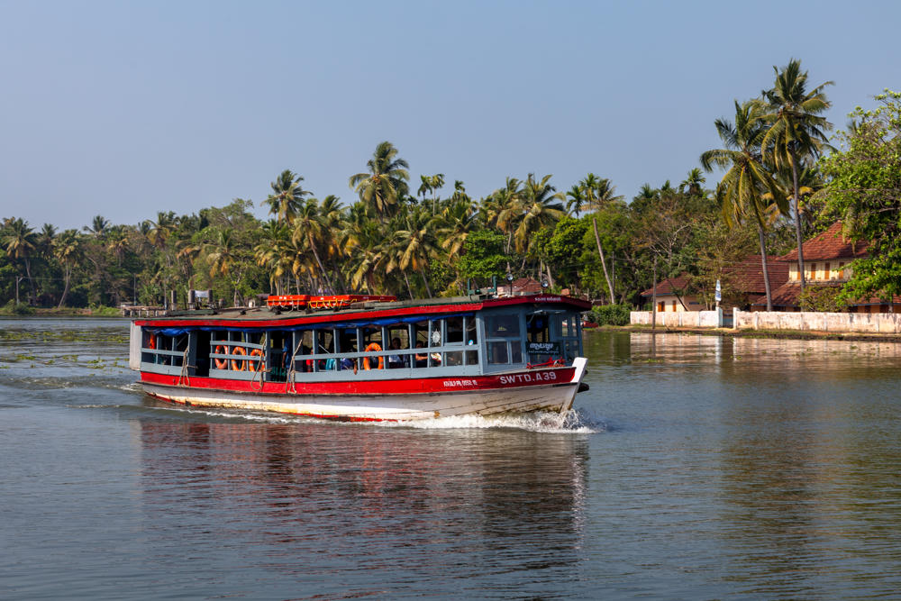 A red and white passenger ferry marked SWTD.A39 carrying people cruises along a calm river, leaving a wake behind it. The far bank is lined with dense palm trees and tropical greenery, with a tiled-roof building visible under a hazy blue sky.