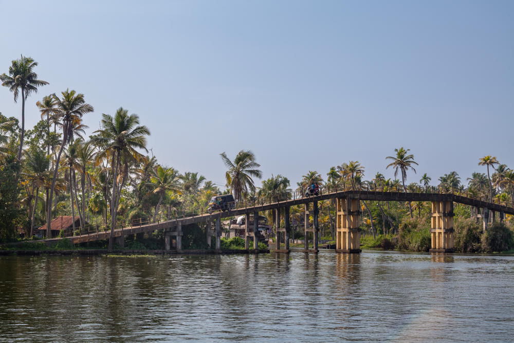 A high concrete road bridge on tall pillars spans the Pamba River, crossed by an auto-rickshaw and a motorcyclist. Dense coconut palms and small houses line the far bank under a bright, hazy white sky, with calm water in the foreground.