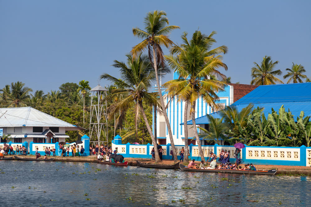 A waterside village along the Pamba River in Kerala, with people crowded along a blue-and-white boundary wall and boarding narrow wooden canoes on the water. Tall coconut palms rise above white and blue buildings, including one with a bright blue metal roof and a metal water tower, under a hazy pale sky.