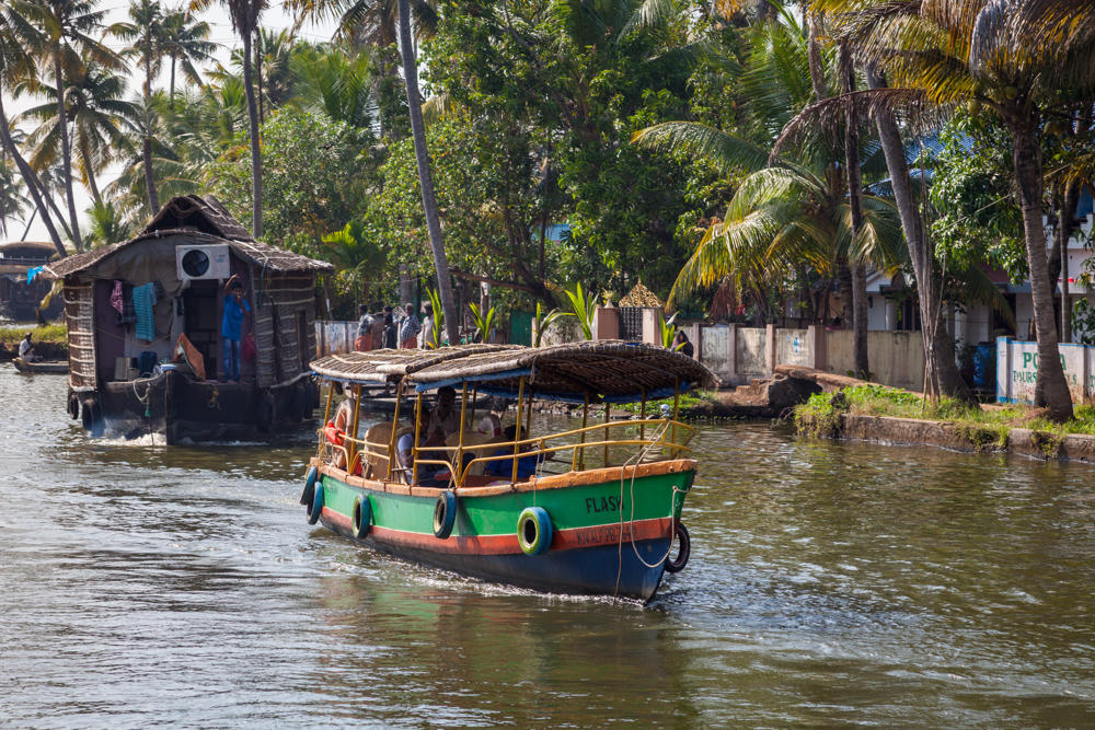 A green and blue passenger boat named “Flash” with a thatched and woven roof carries people along a palm-lined waterway in the Kerala backwaters, with a thatched-roof houseboat behind it and lush greenery and buildings along the far bank.