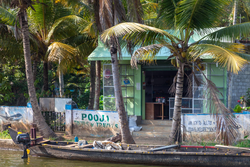 A long wooden canoe with a Suzuki outboard motor sits in a canal in front of a small green-painted shop with an open doorway, its low boundary wall painted with the sign “Pooja Tours & Travels,” all shaded by tall coconut palm trees in the Kerala backwaters.