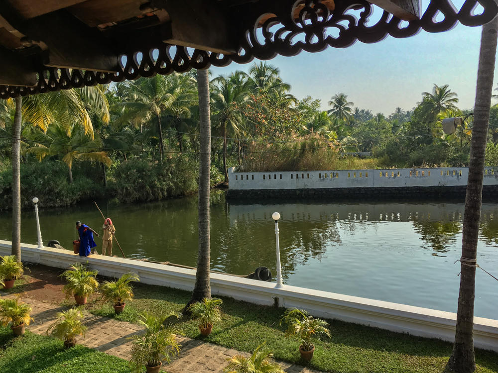 View from beneath an ornately carved dark wooden eave looking out over a calm green backwater lined with tall coconut palms, where two people stand on a white-edged walkway near the water, one holding a long pole beside a boat. Potted palms sit on a paved and grassy garden in the foreground, with a white boundary wall and dense tropical foliage across the water.