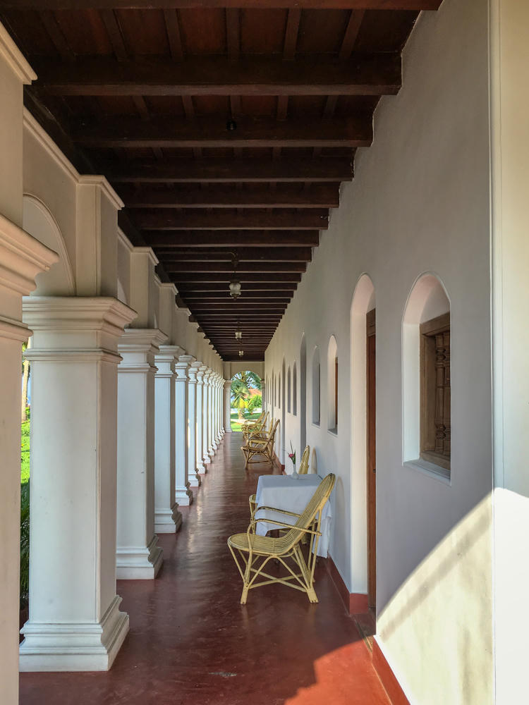 A long open colonnaded verandah with a polished red oxide floor, a row of white plaster columns on the left opening to greenery, and a whitewashed wall with arched niches and carved wooden windows on the right. Yellow cane chairs and cloth-draped tables line the walkway beneath a dark exposed wooden beam ceiling.