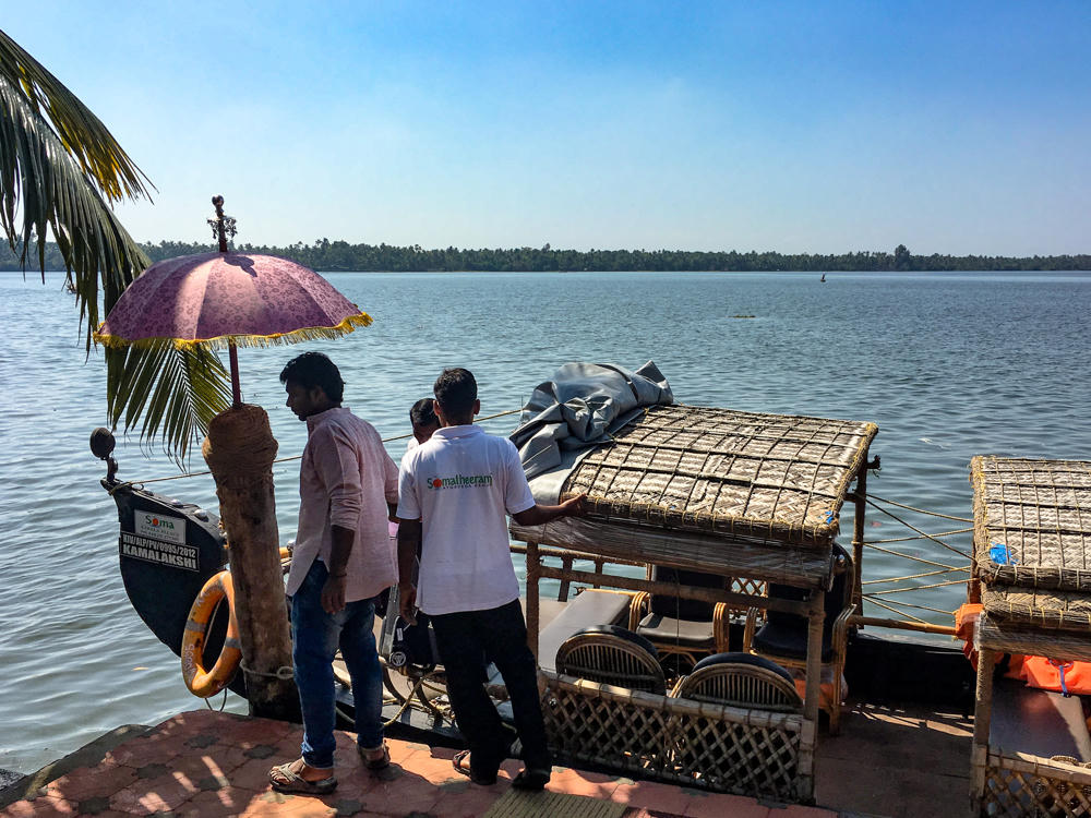 Two men, one in a white Somatheeram staff T-shirt and one in a pink shirt, stand on a tiled waterfront jetty beside a traditional Kerala houseboat with thatched cane roofs, an orange life ring, and a purple decorative umbrella, on a calm backwater under a clear blue sky.