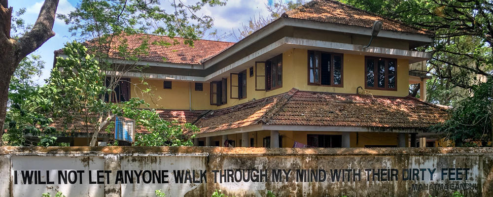 A weathered concrete boundary wall in Fort Kochi painted with a large black quote reading “I WILL NOT LET ANYONE WALK THROUGH MY MIND WITH THEIR DIRTY FEET” attributed to Mahatma Gandhi, with green weeds in the foreground and a tiled-roof building and trees behind the wall.