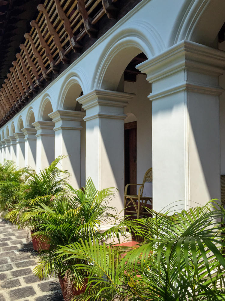 A sunlit colonnade of white plastered archways and square columns runs along a covered walkway, with a wooden slatted eave above and a row of potted areca palms lining the brick-paved floor. A cane armchair sits in the shaded corridor beyond one of the arches.