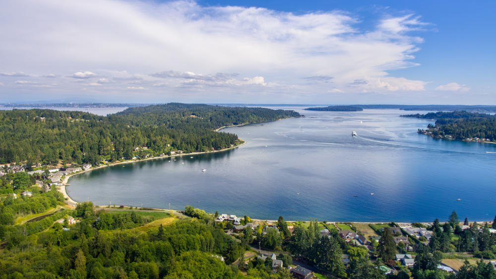 An aerial view of a forested inlet on Bainbridge Island, with a curving bay ringed by evergreen trees and scattered waterfront homes, small boats and a ferry moving across the blue water under a broad sky streaked with wispy clouds.
