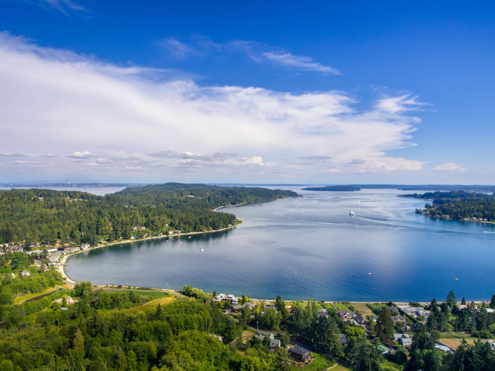 An aerial view of a forested inlet on Bainbridge Island, with a curving bay ringed by evergreen trees and scattered waterfront homes, small boats and a ferry moving across the blue water under a broad sky streaked with wispy clouds.