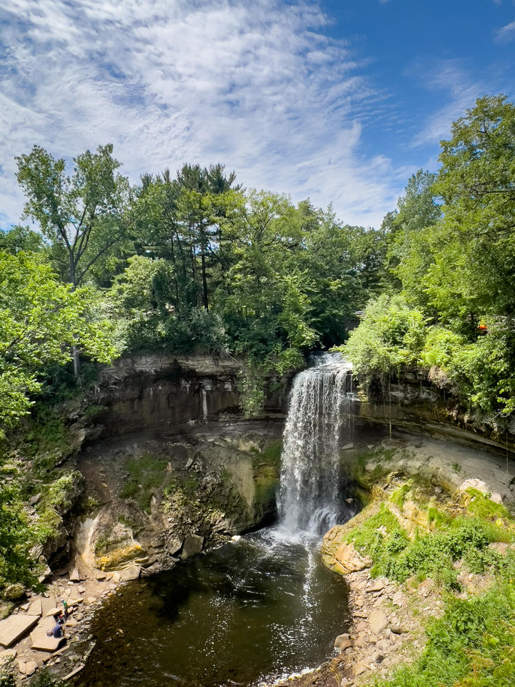 A waterfall spilling over a rocky limestone cliff into a dark pool in a wooded gorge, framed by lush green summer foliage under a bright blue sky with wispy clouds. A couple of small figures stand on the rocks at lower left, giving scale to the scene.