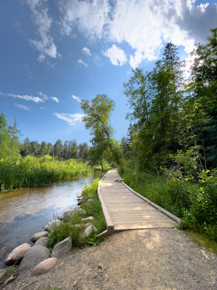 A wooden boardwalk trail curves alongside a clear, gently flowing stream at the Mississippi Headwaters in Itasca State Park, flanked by lush green grasses, reeds, and rounded boulders, with tall pines and leafy trees under a bright blue sky dotted with dramatic white clouds.