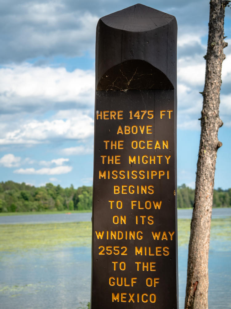 A tall brown wooden marker post with gold lettering that reads “Here 1475 ft above the ocean the mighty Mississippi begins to flow on its winding way 2552 miles to the Gulf of Mexico,” standing beside a lake with lily pads under a partly cloudy sky at the Mississippi Headwaters in Itasca State Park.