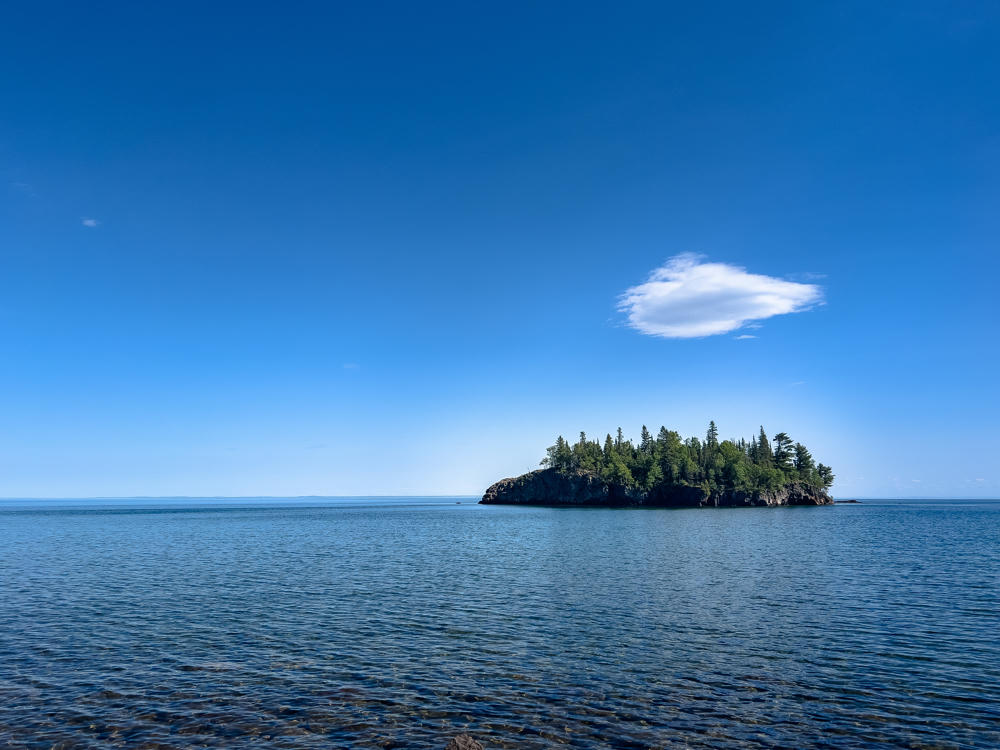 A small rocky island topped with a dense stand of evergreen trees sits in the calm blue waters of a large lake under a clear sky with a single puffy white cloud. Gentle ripples cover the foreground water, with the distant shoreline barely visible on the horizon.