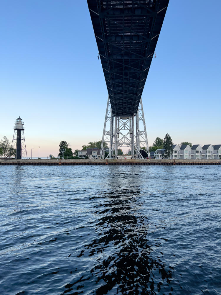 View from the water looking up at the underside of the steel Aerial Lift Bridge in Duluth, Minnesota, with a tall lighthouse on the shore to the left and a row of white townhouses along the waterfront to the right beneath a clear evening sky.