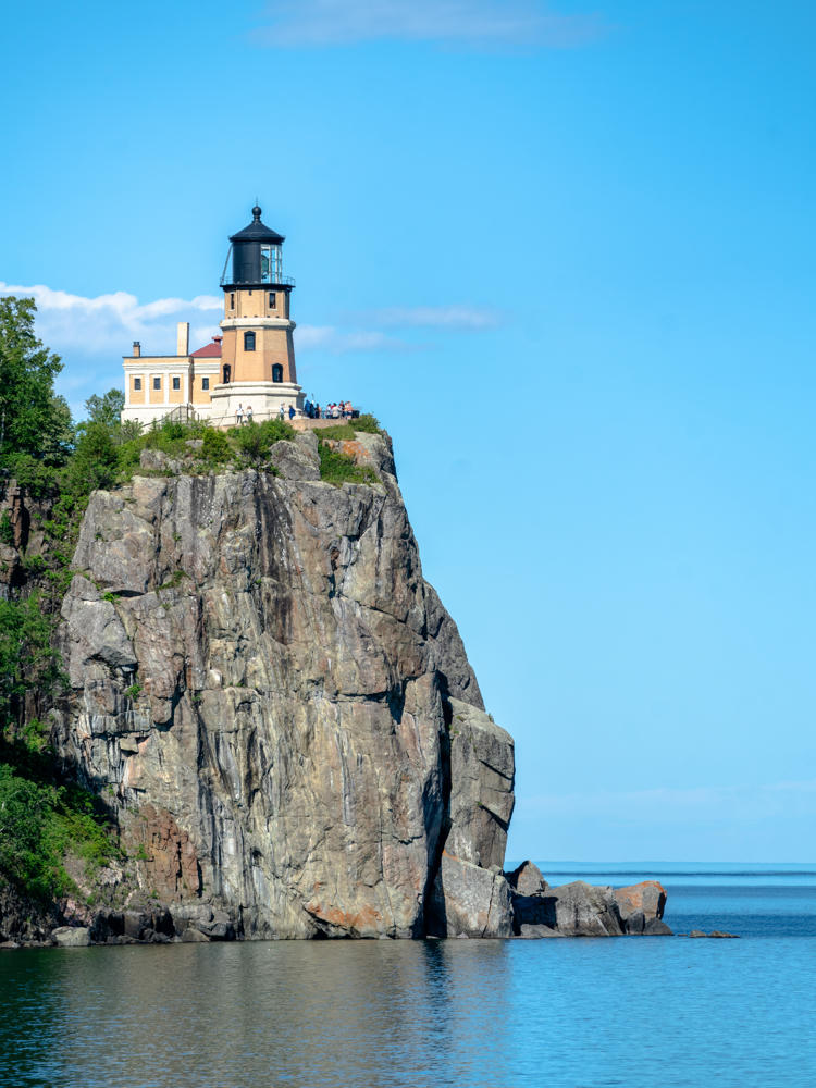Split Rock Lighthouse, a brick tower with a black lantern room, perched atop a steep rocky cliff rising from the blue water of Lake Superior, with visitors gathered on the overlook beside the adjacent keeper’s building under a clear blue sky.