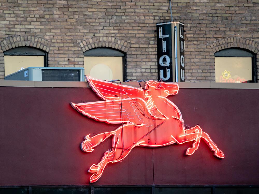 A glowing red neon sign of a winged flying horse (Pegasus) mounted on the dark maroon facade of a brick building, with a vertical neon “LIQUOR” sign partially visible above on the tan brick upper story.