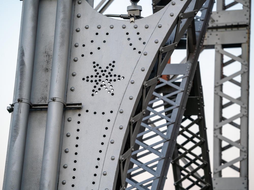 Close-up of a riveted gray steel beam on the Aerial Lift Bridge, with a decorative star-and-lines pattern formed by punched holes in the metal plate, set against latticework girders and a pale sky.