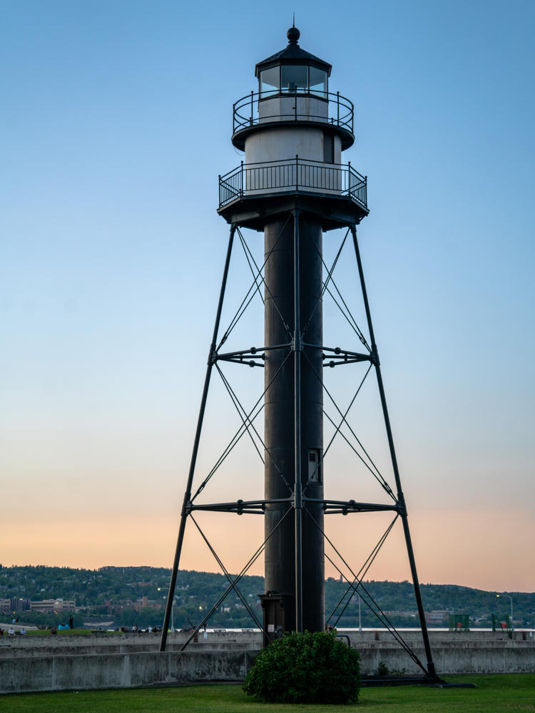 A tall metal skeletal lighthouse with a cylindrical central tower and a white lantern room silhouetted against a soft pale blue and peach twilight sky, standing on a grassy area near a concrete pier with a low city skyline and wooded hills on the horizon.