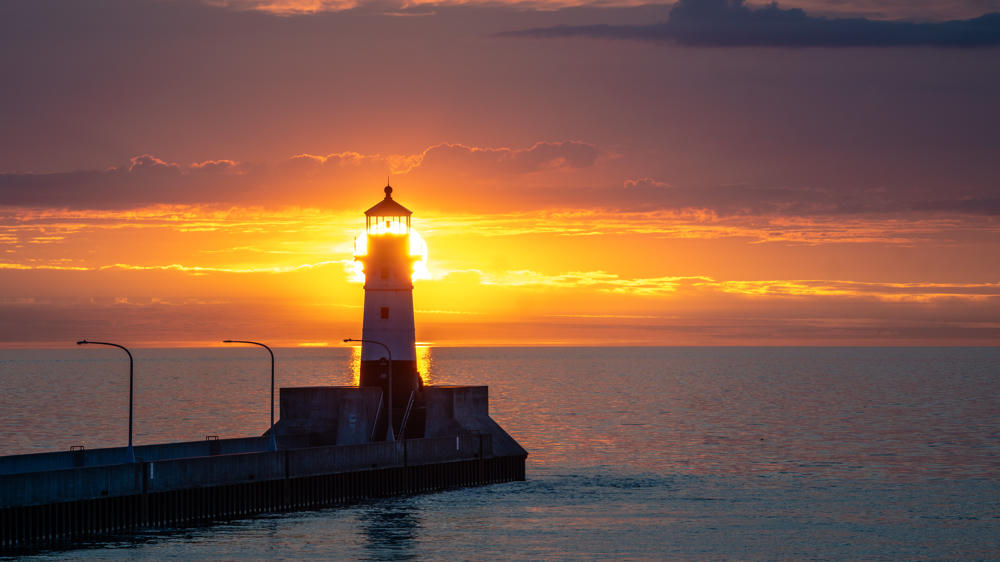 A lighthouse at the end of a long concrete pier silhouetted against a vivid orange sunrise over Lake Superior, with the sun’s glow radiating from behind the tower and lampposts lining the walkway.