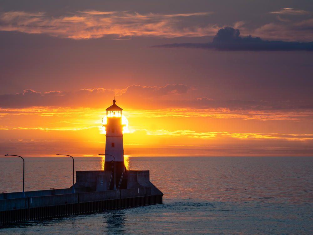 A lighthouse at the end of a long concrete pier silhouetted against a vivid orange sunrise over Lake Superior, with the sun’s glow radiating from behind the tower and lampposts lining the walkway.