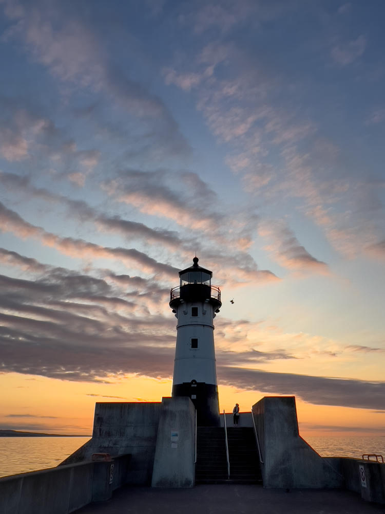 A white lighthouse with a black lantern room stands atop a concrete pier at dawn, silhouetted against a dramatic sky streaked with pink and orange clouds over calm water. A lone figure stands on the pier near the base of the tower, and a bird flies near the lantern gallery.