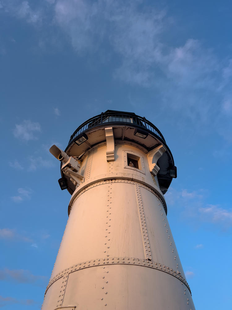 Looking up at the riveted white steel tower of the Duluth Harbor North Pier Lighthouse, topped by a black lantern gallery and railing, set against a blue sky streaked with wispy clouds in early morning light.