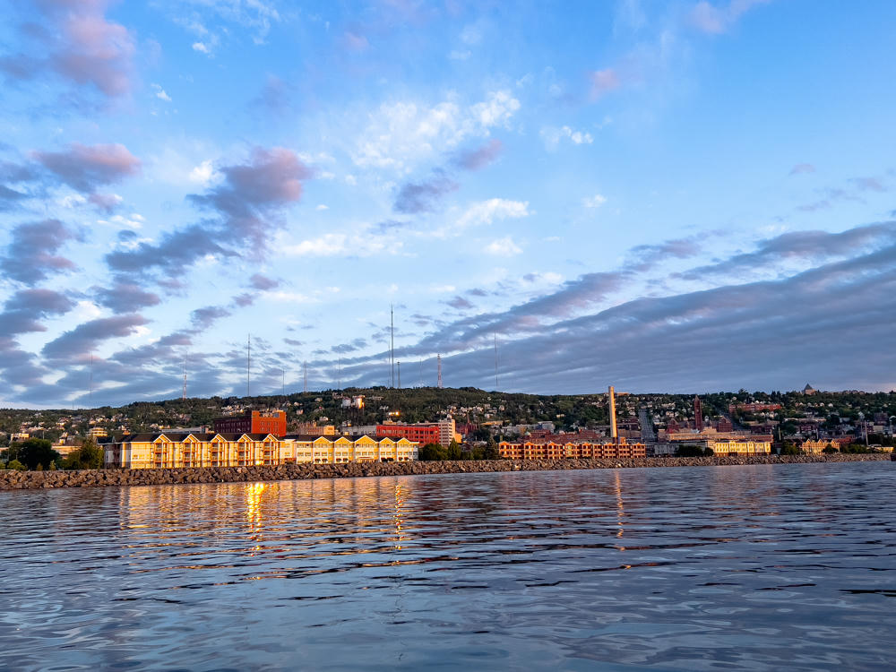 A view across the calm water of Duluth Harbor toward the waterfront at dawn, with warm early light striking rows of buildings and a smokestack along the rocky shore, and a forested hillside dotted with houses and communication towers rising under a partly cloudy sky.