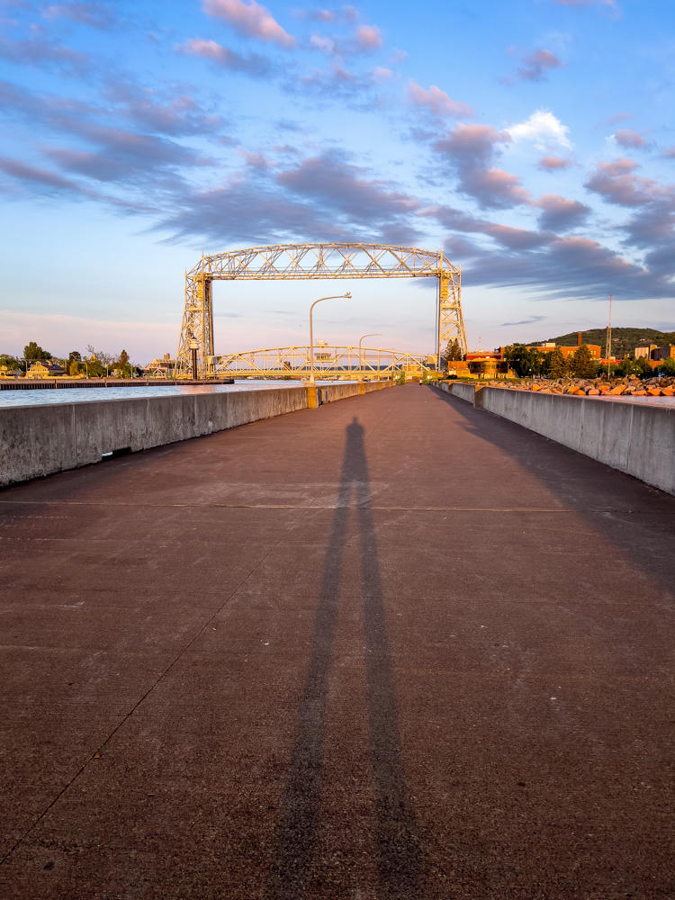 A long concrete pier stretches toward the Aerial Lift Bridge in Duluth, its steel towers lit warm gold by early morning sun, with the photographer’s long shadow cast down the center of the walkway. Blue sky with scattered clouds sits above the harbor, waterfront buildings, and a wooded hillside in the distance.